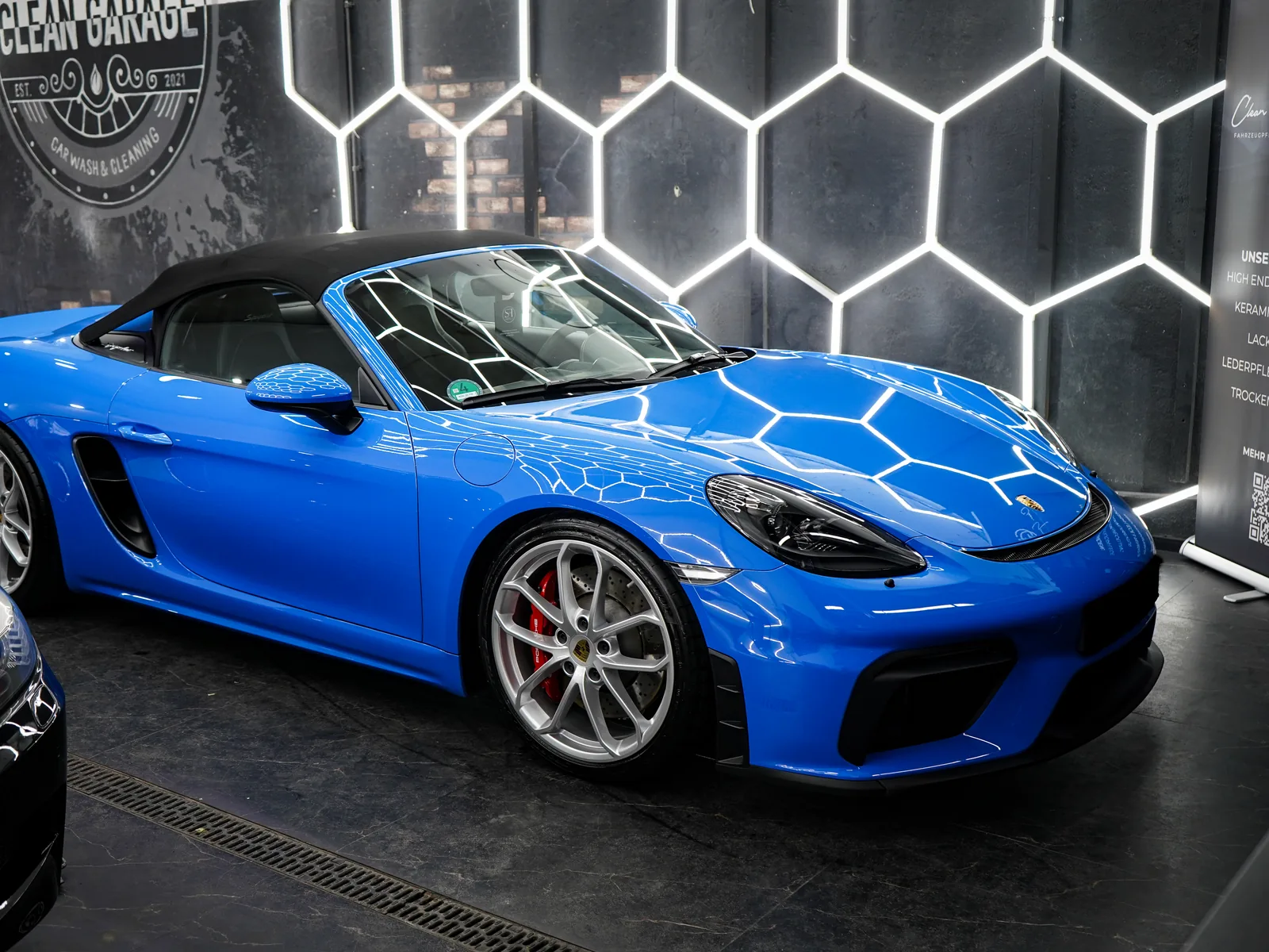 Blauer Porsche 718 Spyder in der Halle von Clean Garage Rheingau, Hex-LED-Decke spiegelt sich im Lack